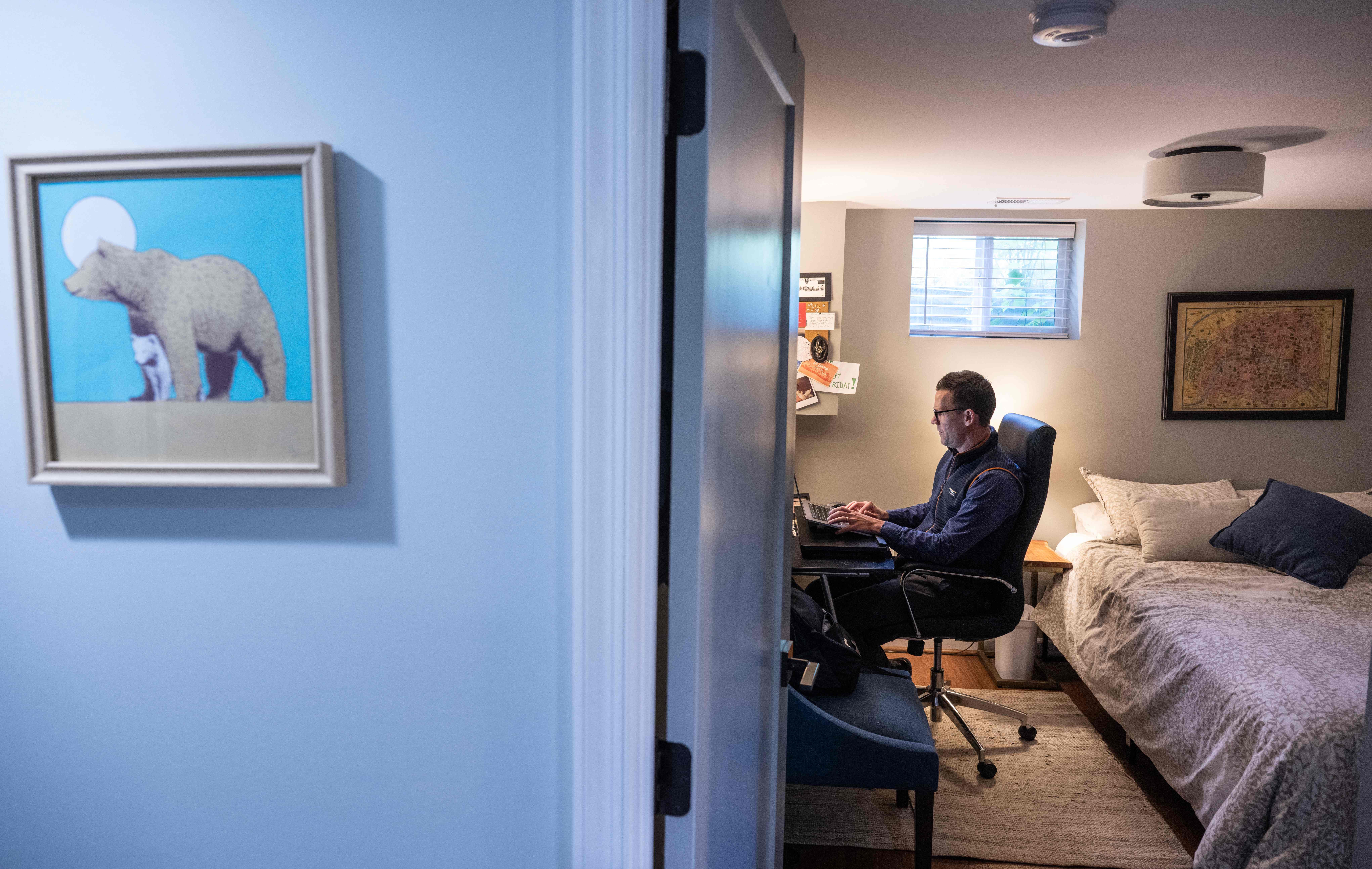 A man sits at an office chair and desk in a bedroom.