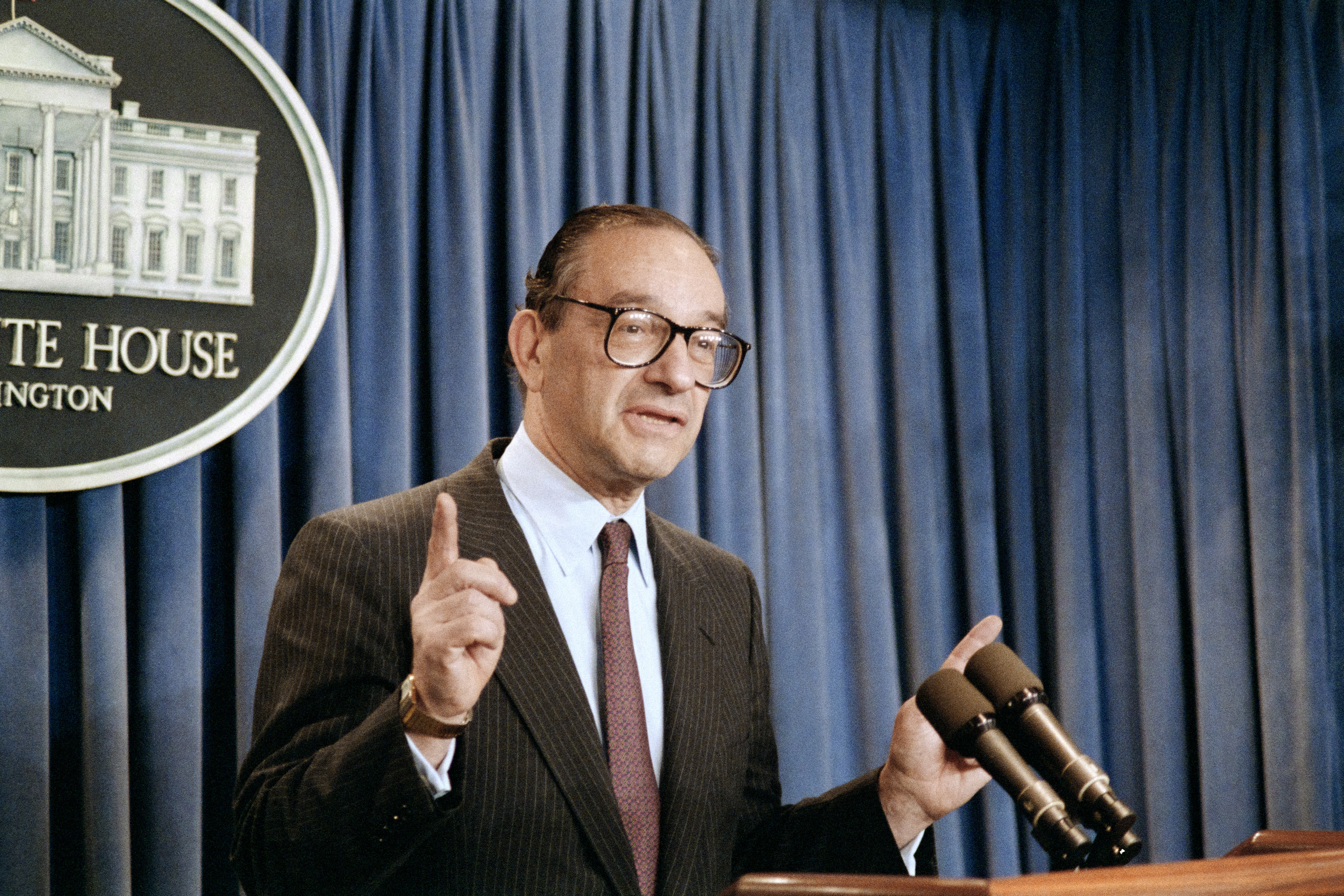 Alan Greenspan, a balding man in glasses, addresses the press in an archival photo