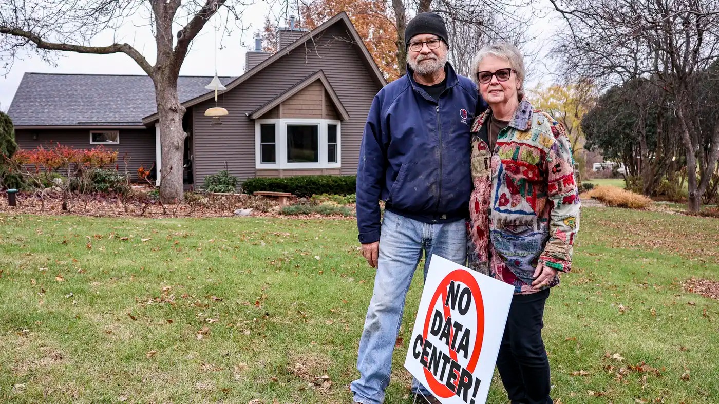A couple stands in front of their home with a sign reading 