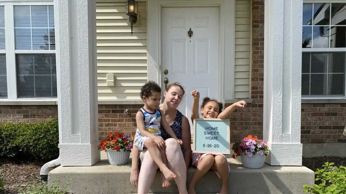 A woman sits on her front stoop of a new home with two children.