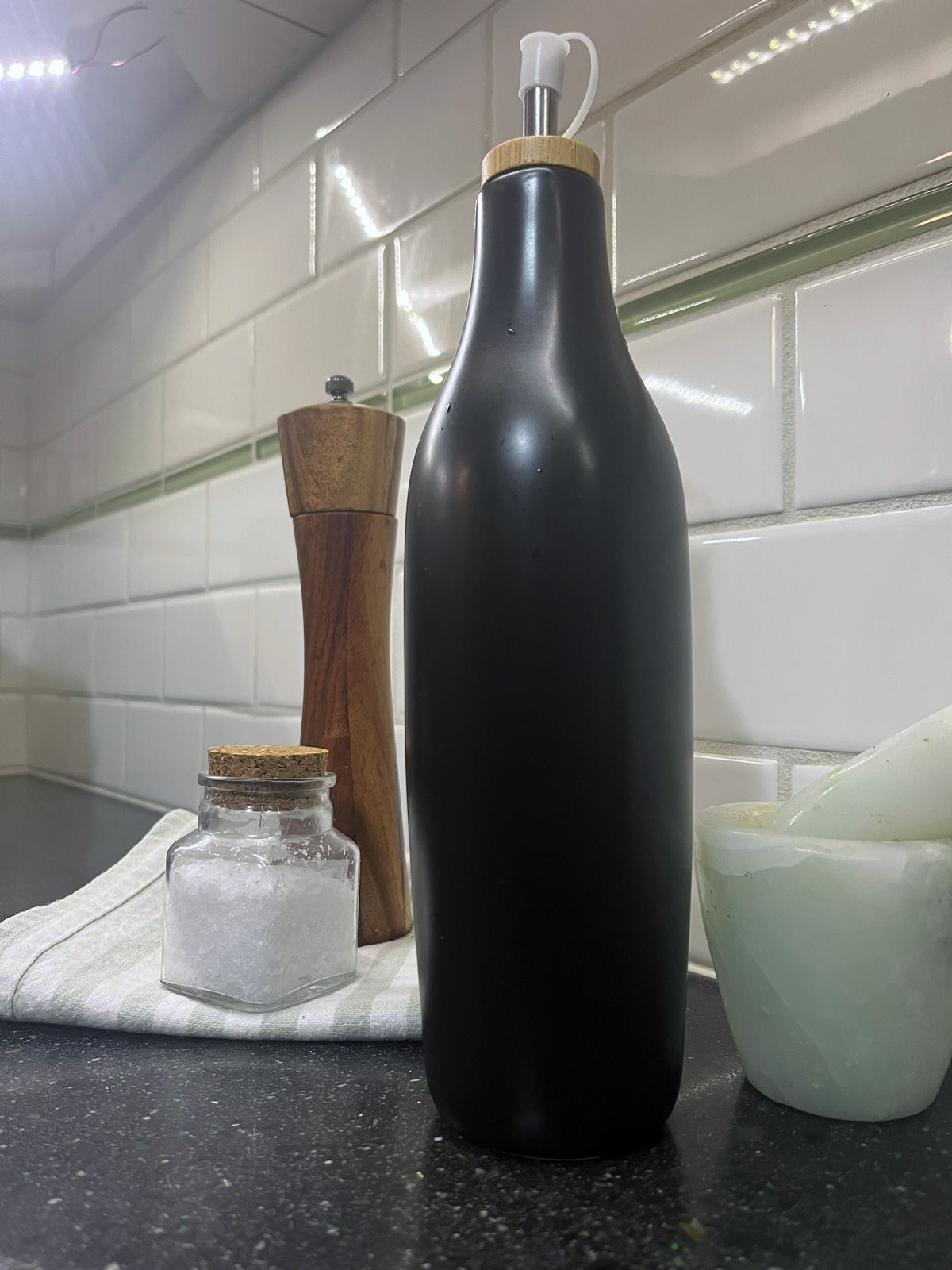 A black bottle of olive oil sits on the counter against white subway tile, next to a pepper grinder and salt cellar.