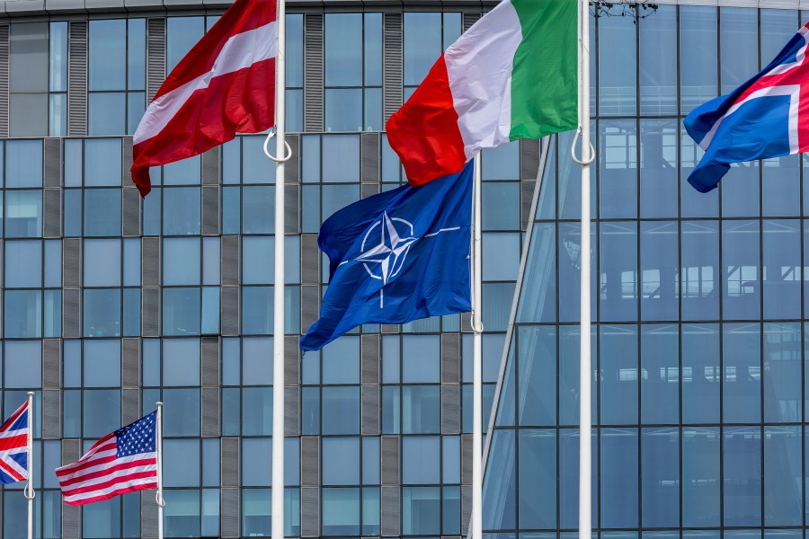 Several national flags on flag poles outside a building flutter in the wind. The NATO flag has a four-point star in the center. 