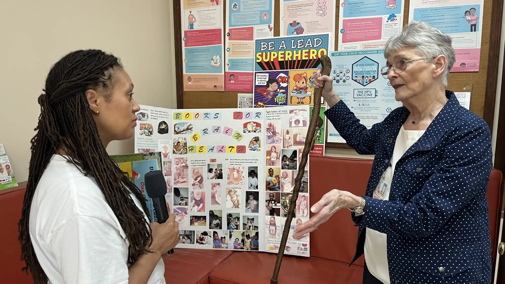 A woman with a microphone speaks with an older woman next to a cork board.