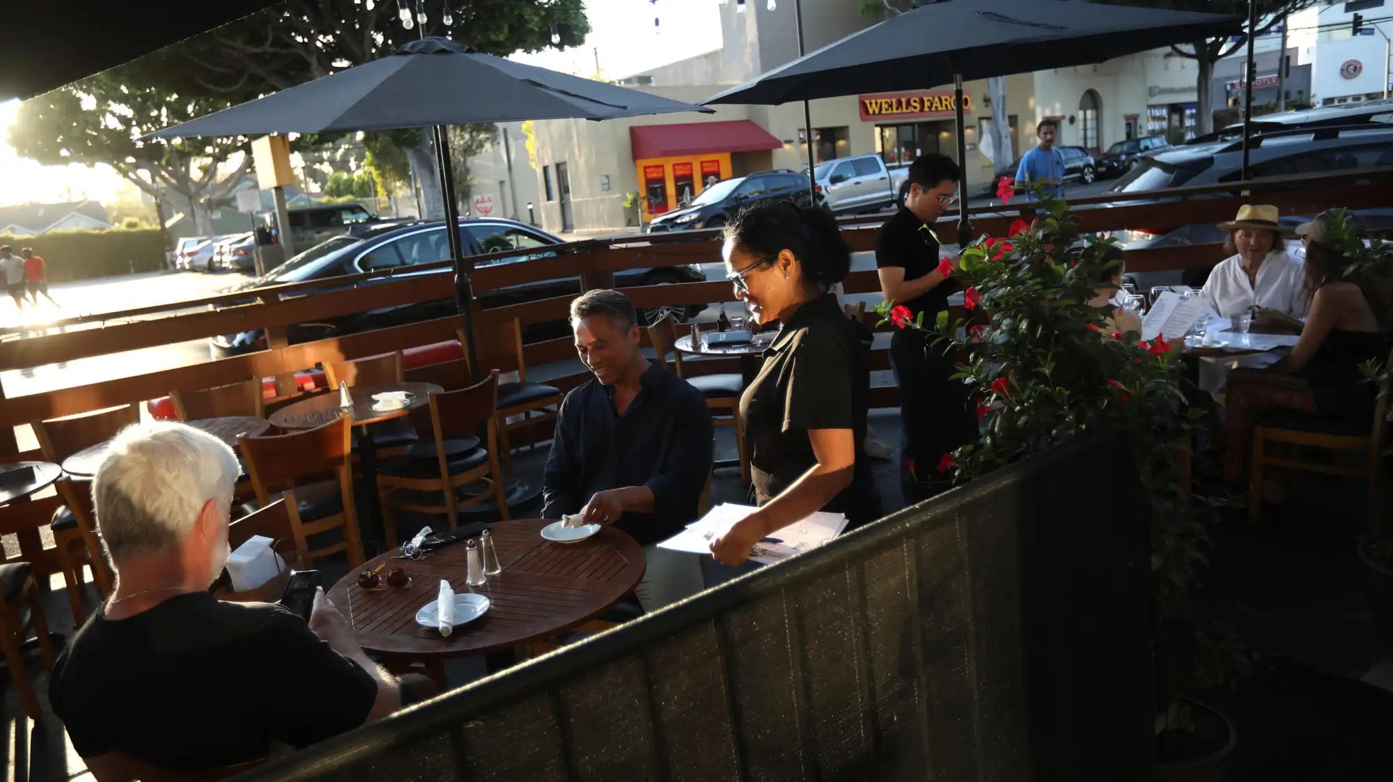 Two people sit at an outdoor restaurant table while a waitress takes their order.
