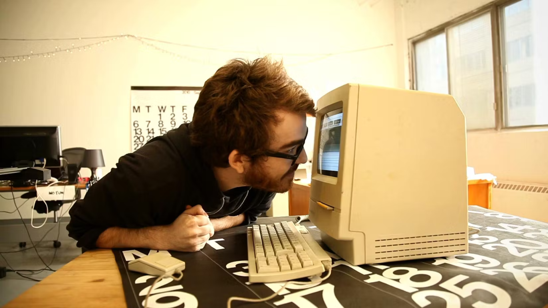 A man sits at a table. He stares at a desktop computer, his face only inches from the screen. 