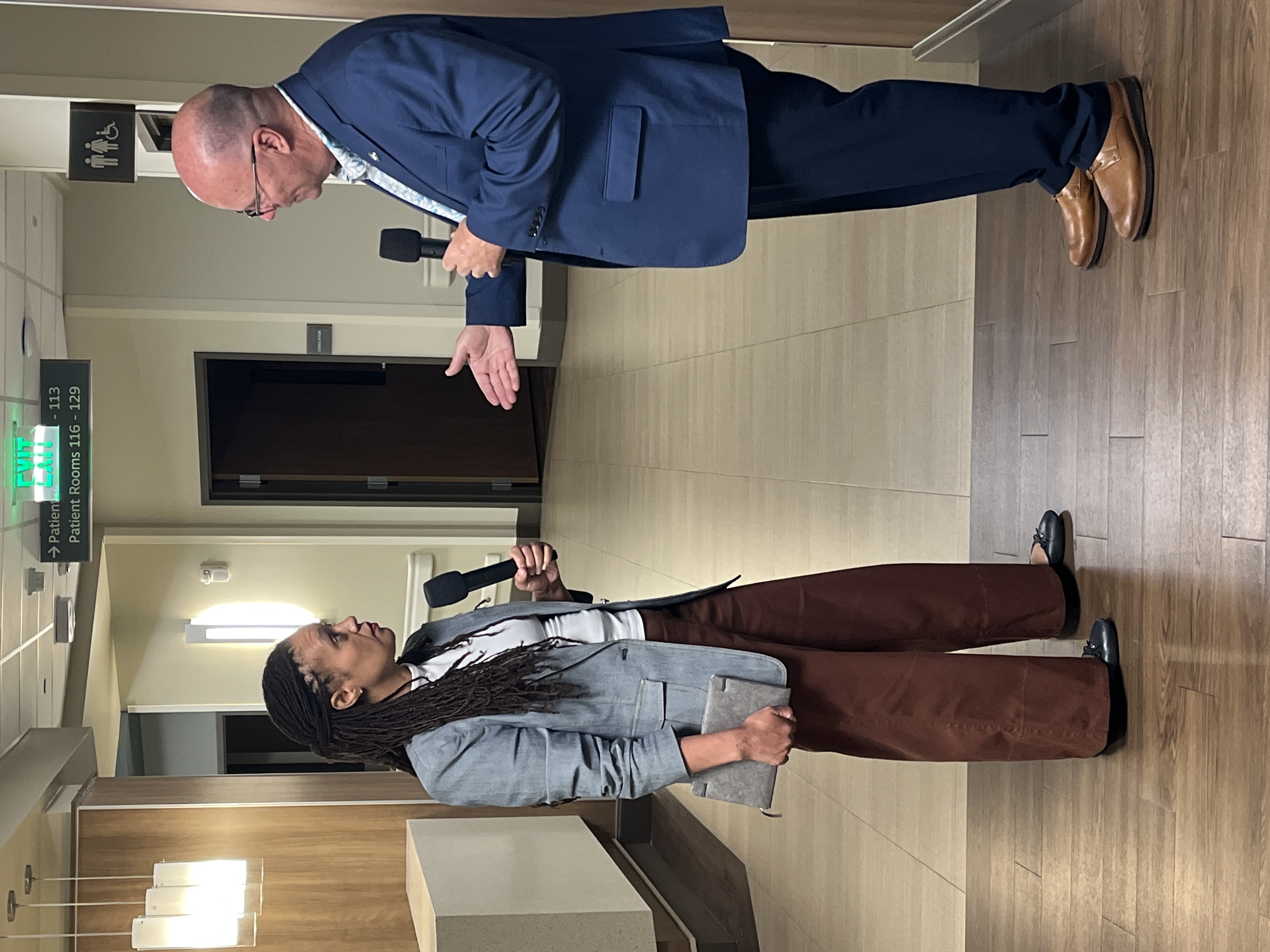 A woman in a blazer and slacks talks to a man in a suit in an empty hospital hallway.