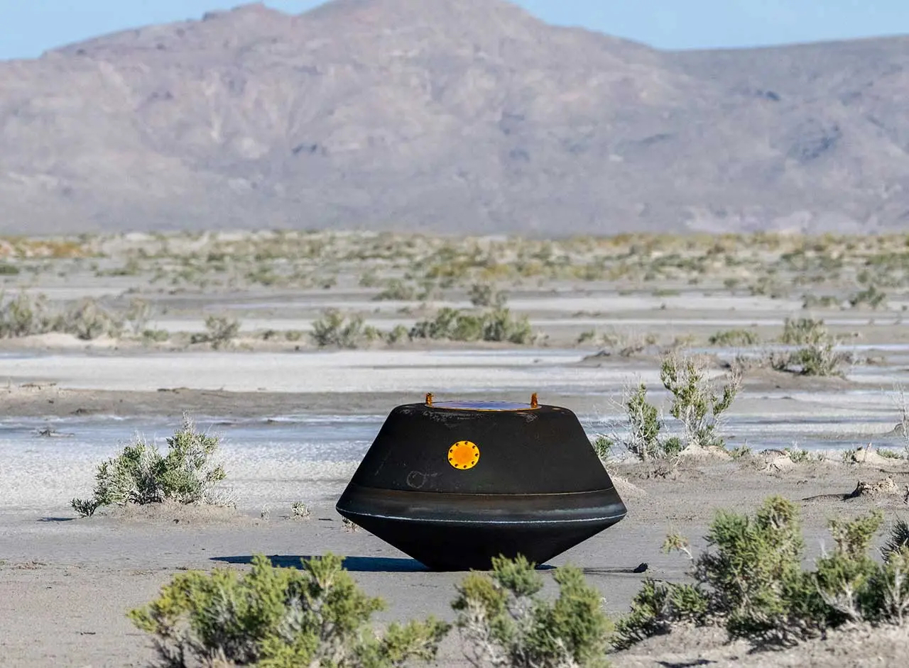 A small capsule sits in the arid Utah desert after returning from space. Shrubs are nearby with mountains in the distance.