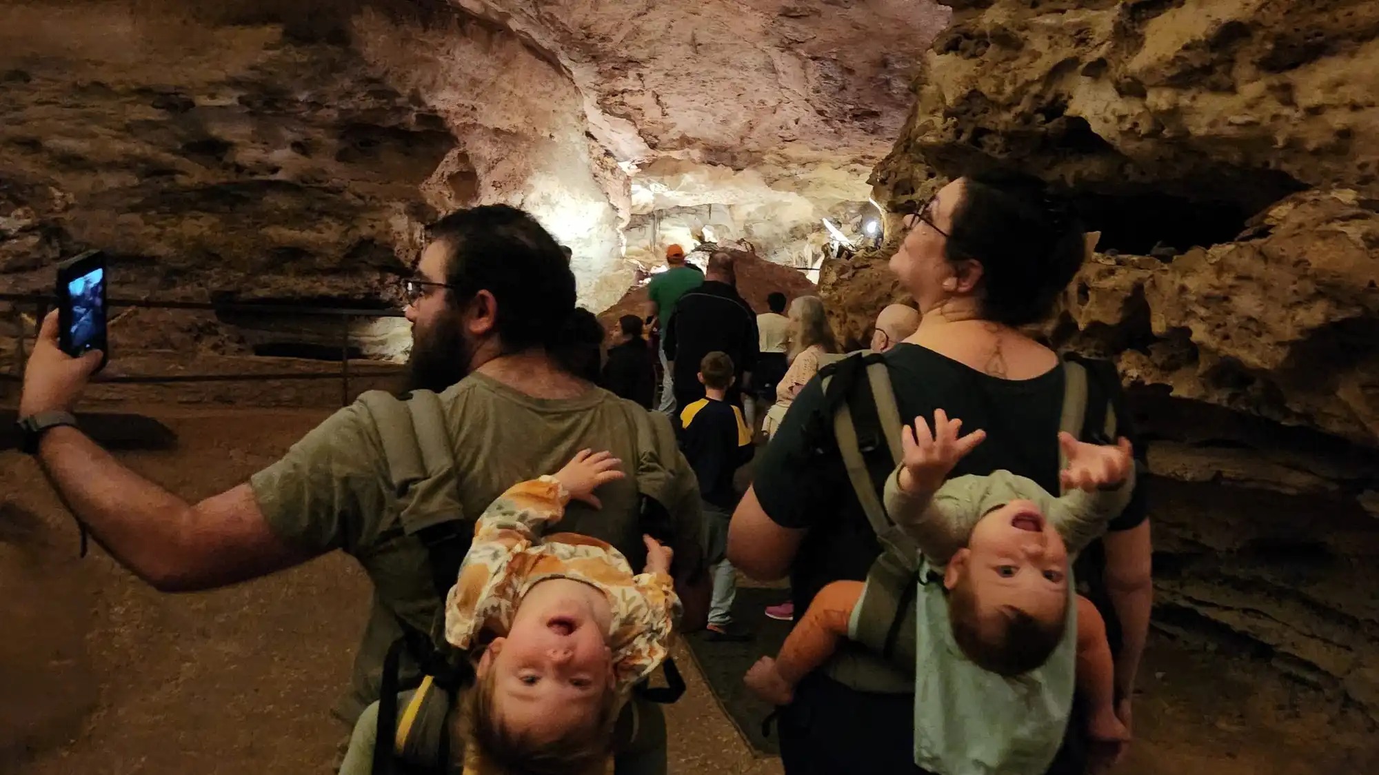 Two people walk through a cave on a tour. They each have a child in a carrier on their back, and both toddlers are leaning back to look at the camera upside-down.