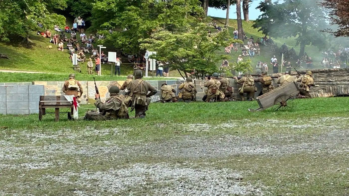 WWII reenacters in a field, watched by regular people sitting on a hillside.