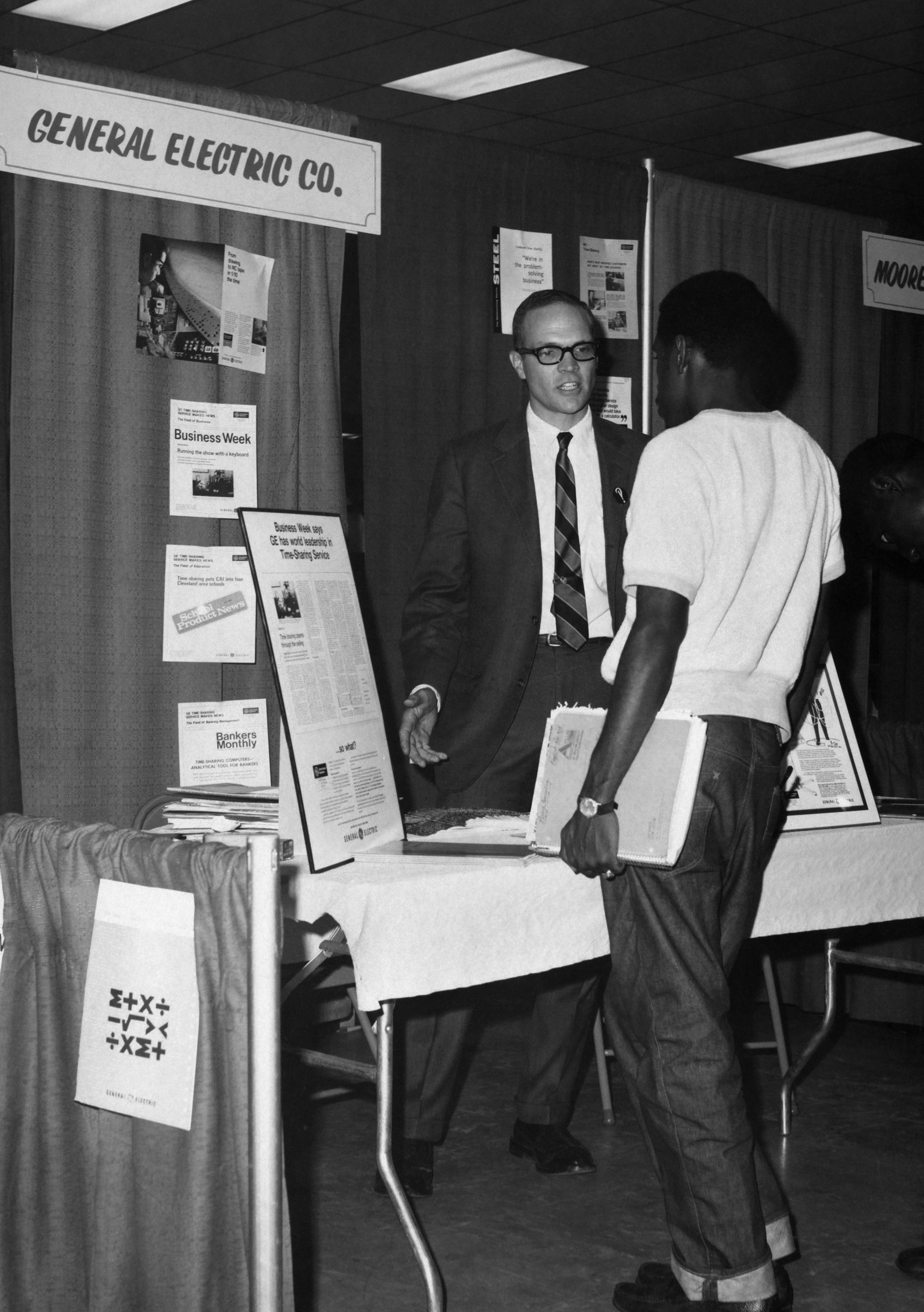 Two men talk at the General Electric booth during a college career fair in the middle of the 20th century.