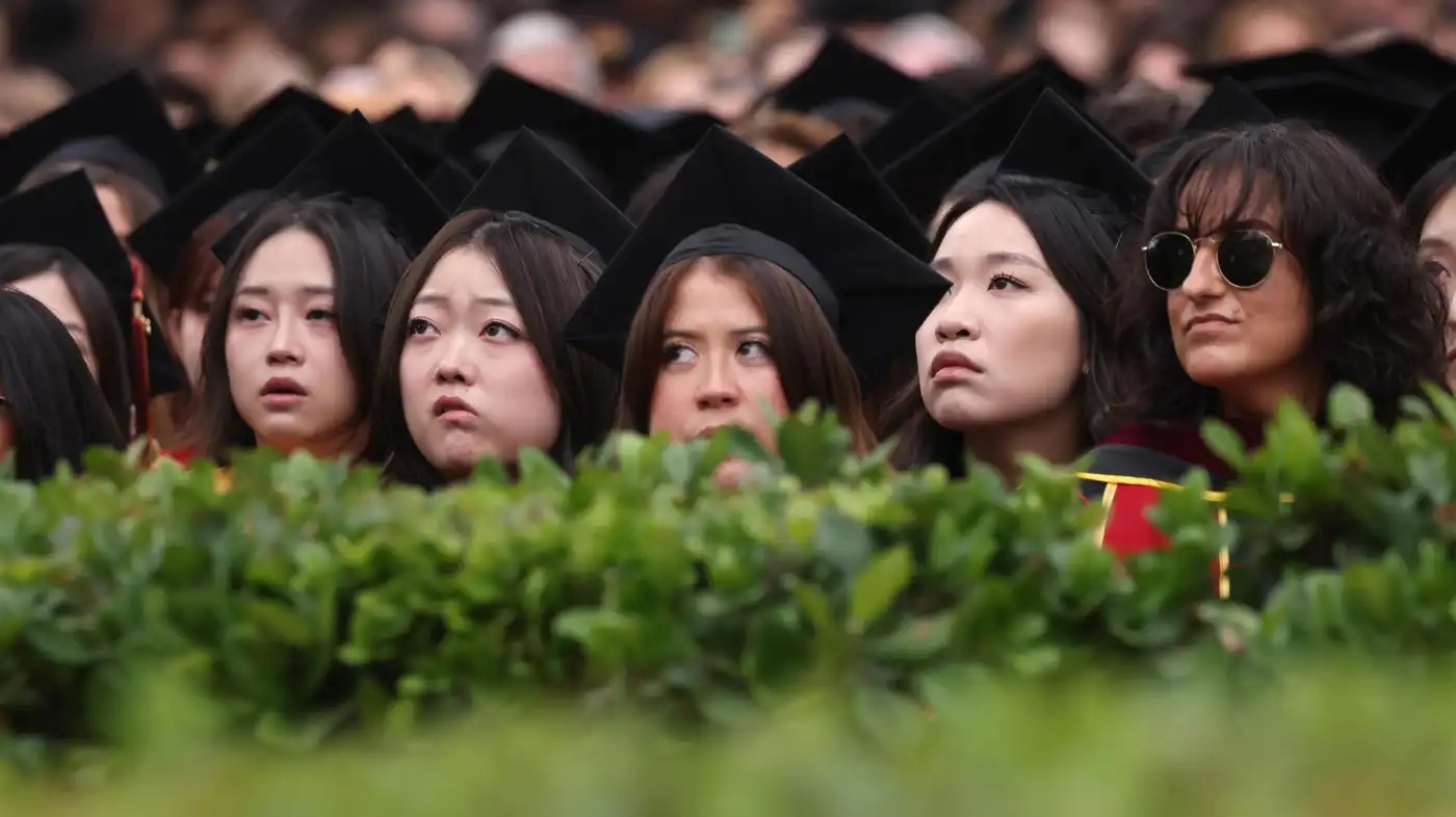 Young women peer over a hedge at a college graduation ceremony. 