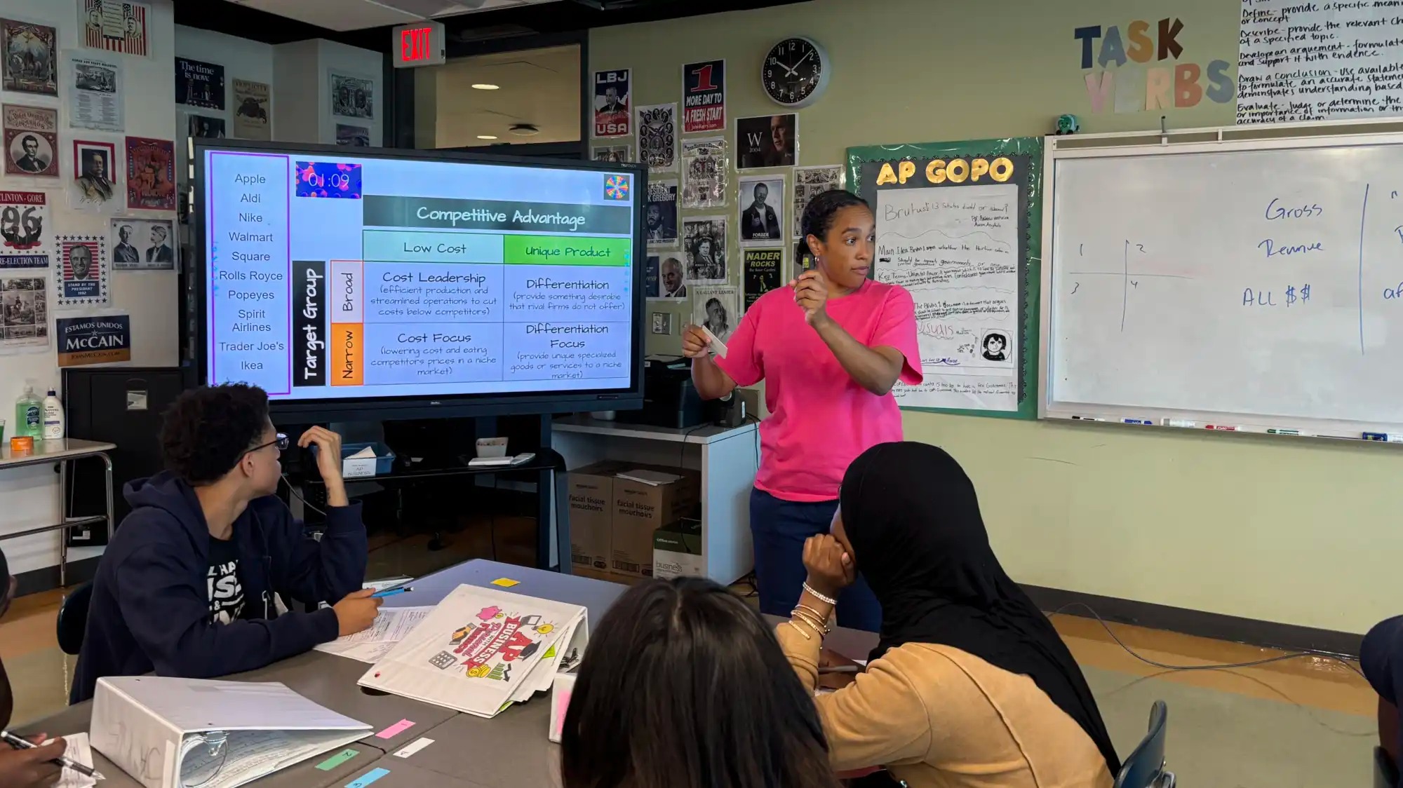 A woman in a bright pink shirt stands in front of a classroom as she gestures to a big screen with information for a lesson in competitive advantage. Three students listen to her speak.